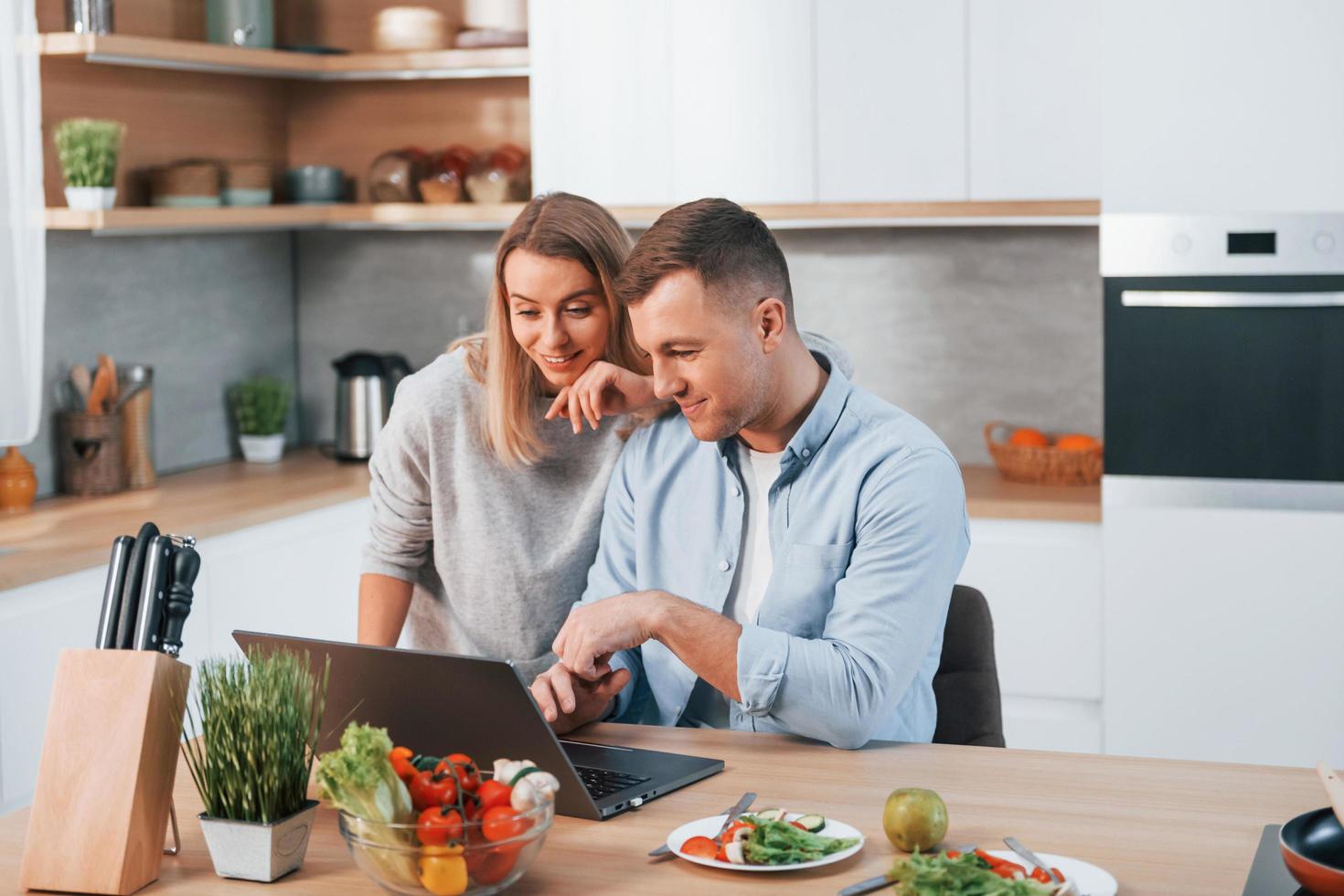 regardant un ordinateur portable. couple préparant la nourriture à la maison dans la cuisine moderne photo