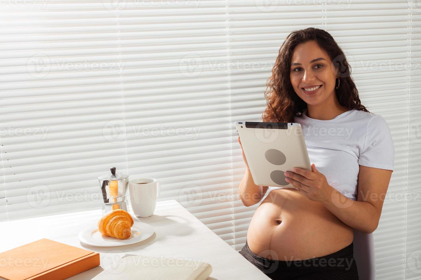 femme enceinte hispanique utilisant une tablette numérique pendant le petit-déjeuner. technologie, grossesse et congé de maternité photo