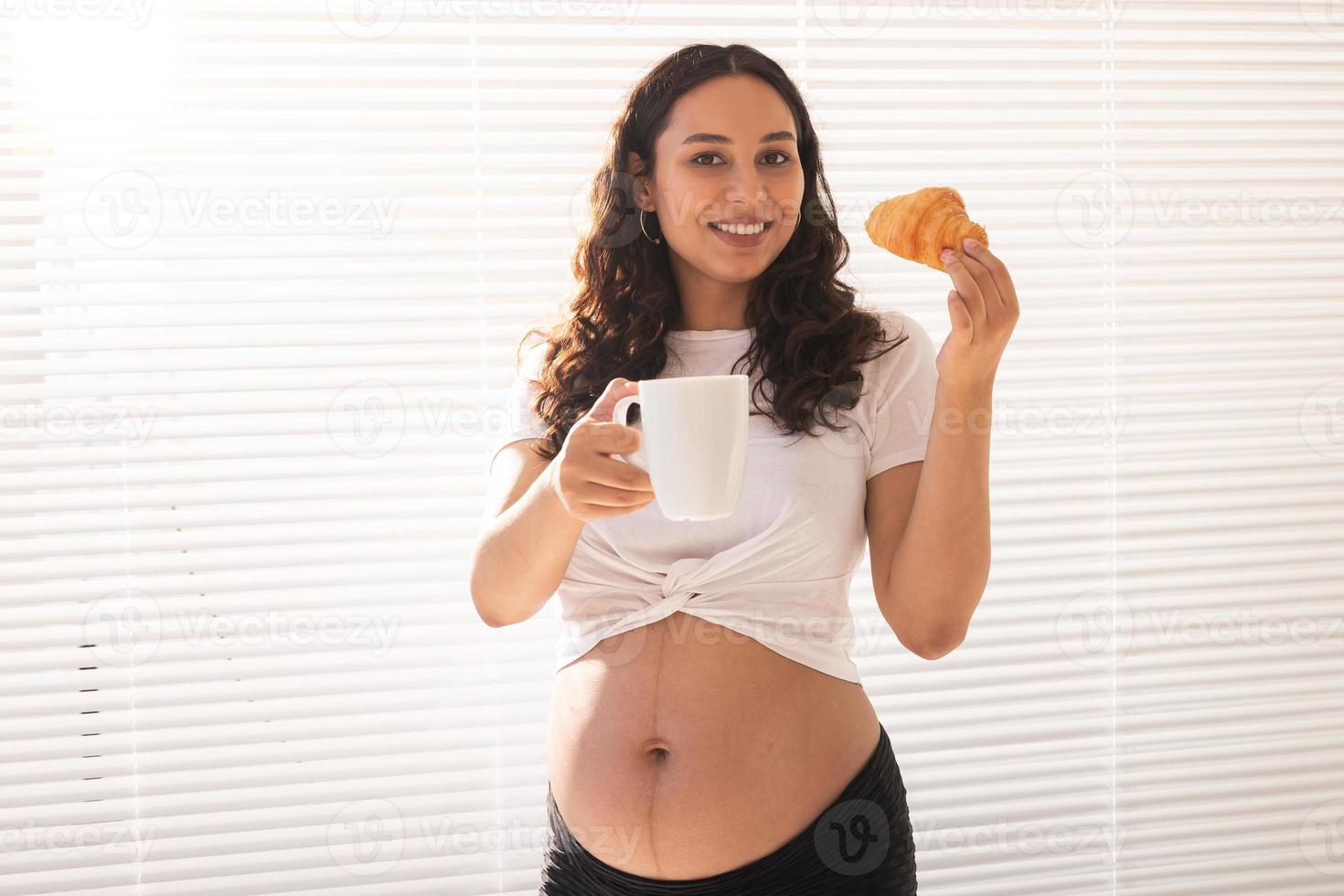belle femme enceinte tenant un croissant et une tasse de café dans ses mains pendant le petit déjeuner du matin. concept de bonne santé et d'attitude positive en attendant bébé photo