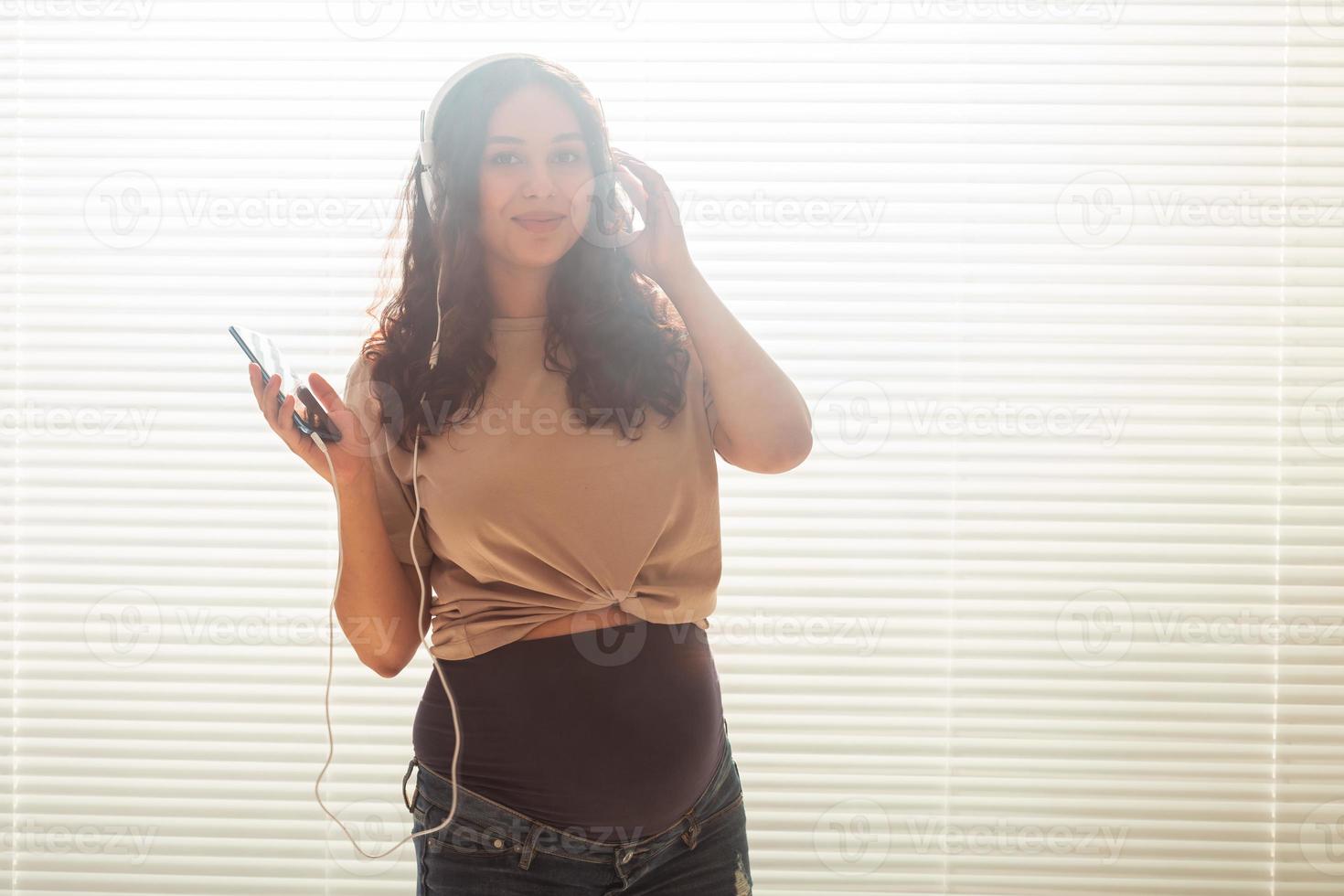 portrait de femme enceinte près de la fenêtre à la maison et écoute de la musique dans des écouteurs avec espace de copie. concept de grossesse et de loisirs. photo