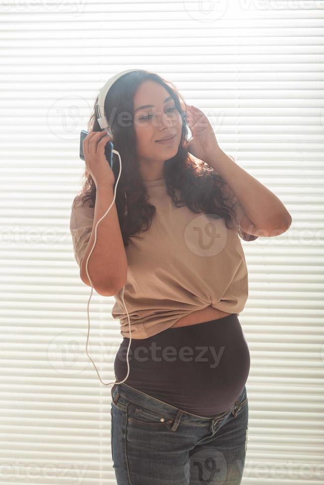 portrait de femme enceinte près de la fenêtre à la maison et écoute de la musique dans les écouteurs. concept de grossesse et de loisirs. photo