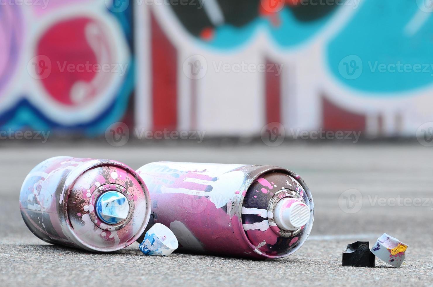 plusieurs bombes aérosols usagées avec de la peinture rose et blanche et des bouchons pour pulvériser de la peinture sous pression se trouvent sur l'asphalte près du mur peint dans des dessins de graffitis colorés photo