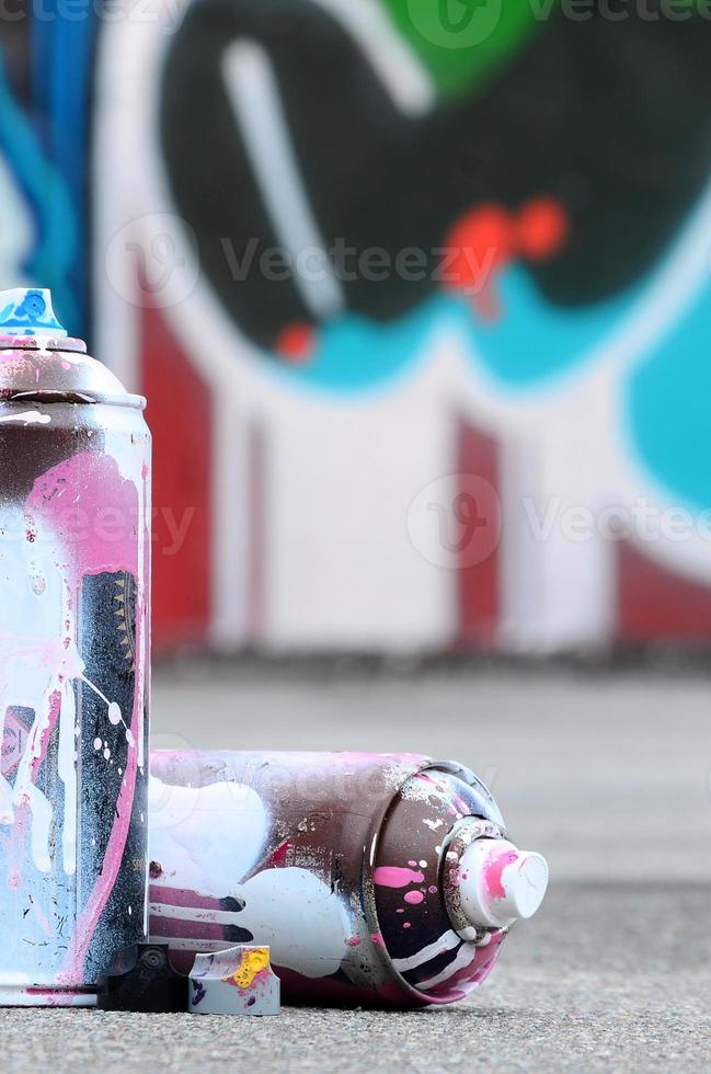 plusieurs bombes aérosols usagées avec de la peinture rose et blanche et des bouchons pour pulvériser de la peinture sous pression se trouvent sur l'asphalte près du mur peint dans des dessins de graffitis colorés photo