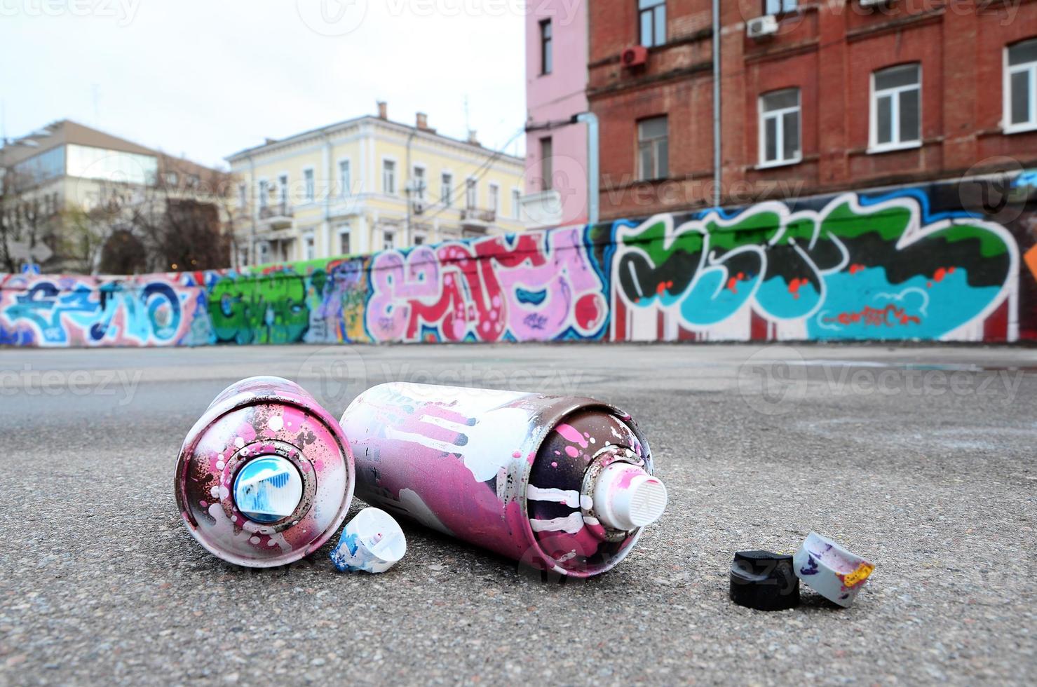 plusieurs bombes aérosols usagées avec de la peinture rose et blanche et des bouchons pour pulvériser de la peinture sous pression se trouvent sur l'asphalte près du mur peint dans des dessins de graffitis colorés photo