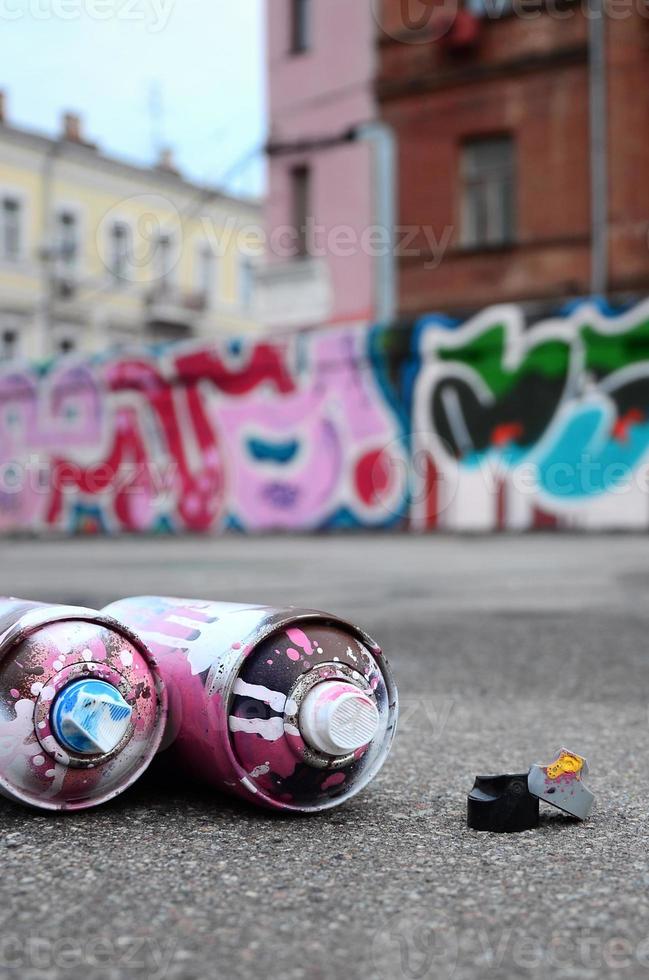 plusieurs bombes aérosols usagées avec de la peinture rose et blanche et des bouchons pour pulvériser de la peinture sous pression se trouvent sur l'asphalte près du mur peint dans des dessins de graffitis colorés photo