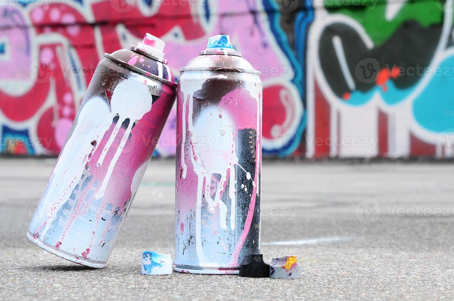 plusieurs bombes aérosols usagées avec de la peinture rose et blanche et des bouchons pour pulvériser de la peinture sous pression se trouvent sur l'asphalte près du mur peint dans des dessins de graffitis colorés photo