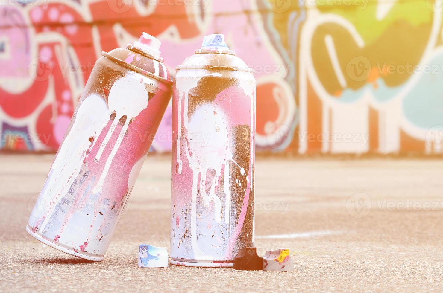 plusieurs bombes aérosols usagées avec de la peinture rose et blanche et des bouchons pour pulvériser de la peinture sous pression se trouvent sur l'asphalte près du mur peint dans des dessins de graffitis colorés photo