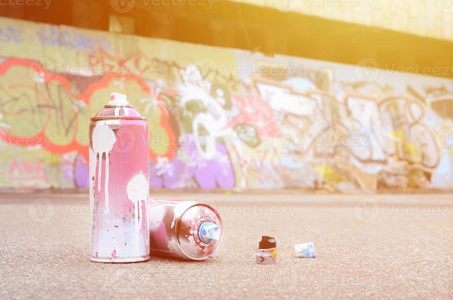 plusieurs bombes aérosols usagées avec de la peinture rose et blanche et des bouchons pour pulvériser de la peinture sous pression se trouvent sur l'asphalte près du mur peint dans des dessins de graffitis colorés photo