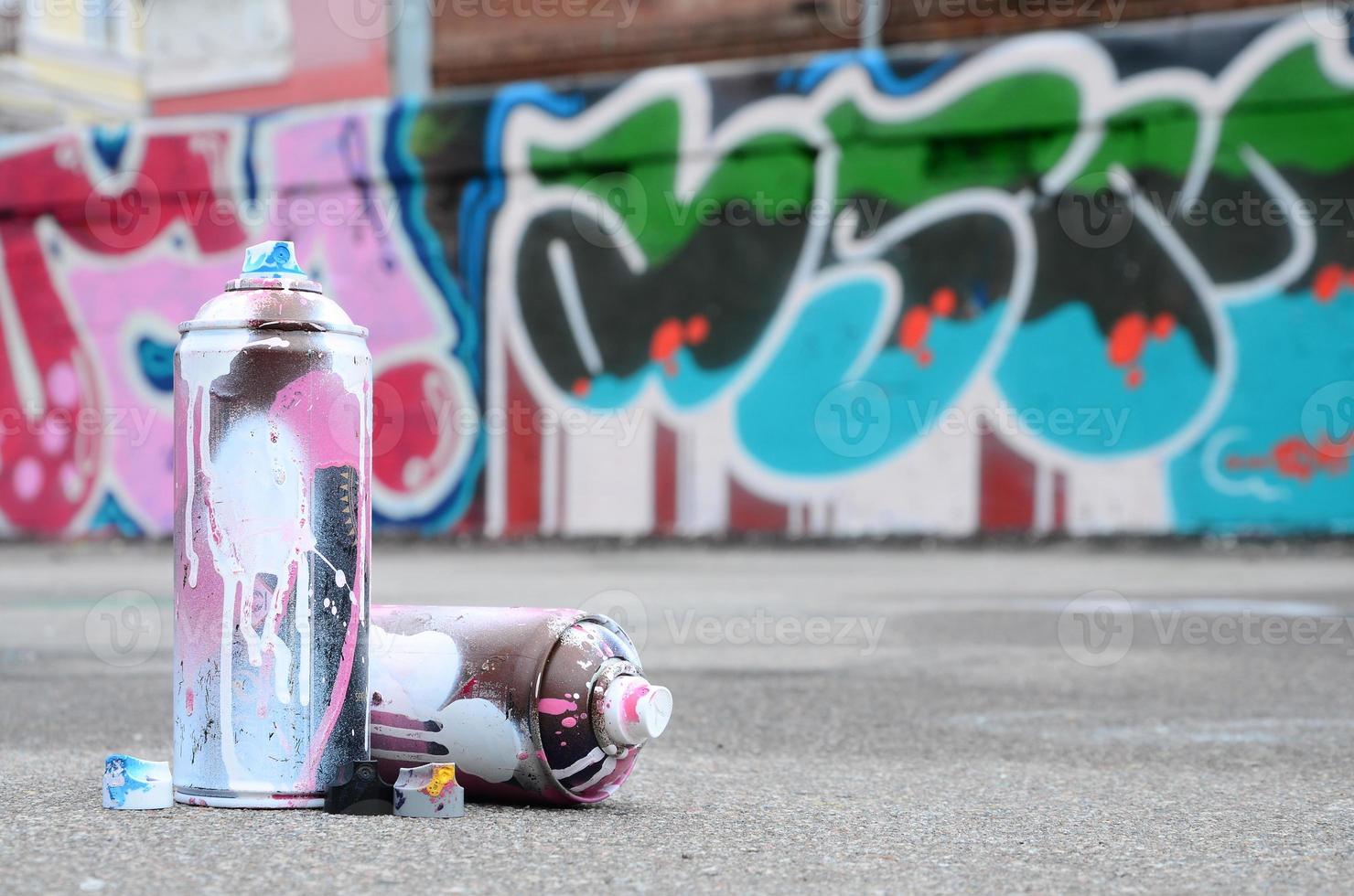 plusieurs bombes aérosols usagées avec de la peinture rose et blanche et des bouchons pour pulvériser de la peinture sous pression se trouvent sur l'asphalte près du mur peint dans des dessins de graffitis colorés photo