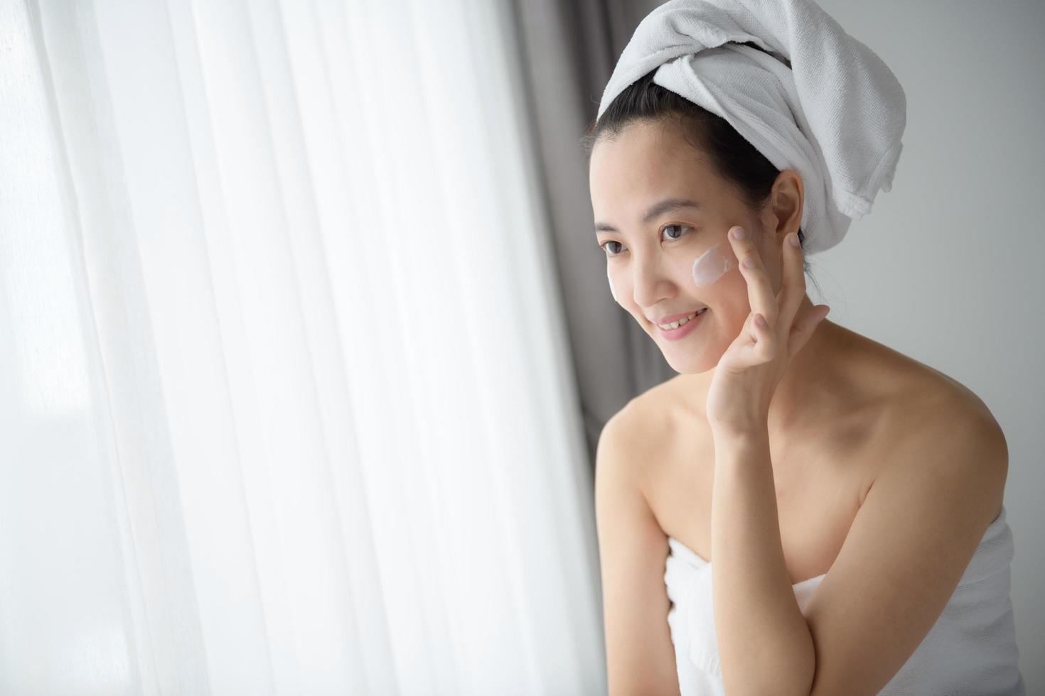 heureuse jeune femme asiatique appliquant des lotions pour le visage tout en portant une serviette et en touchant son visage. maquillage et soins quotidiens photo