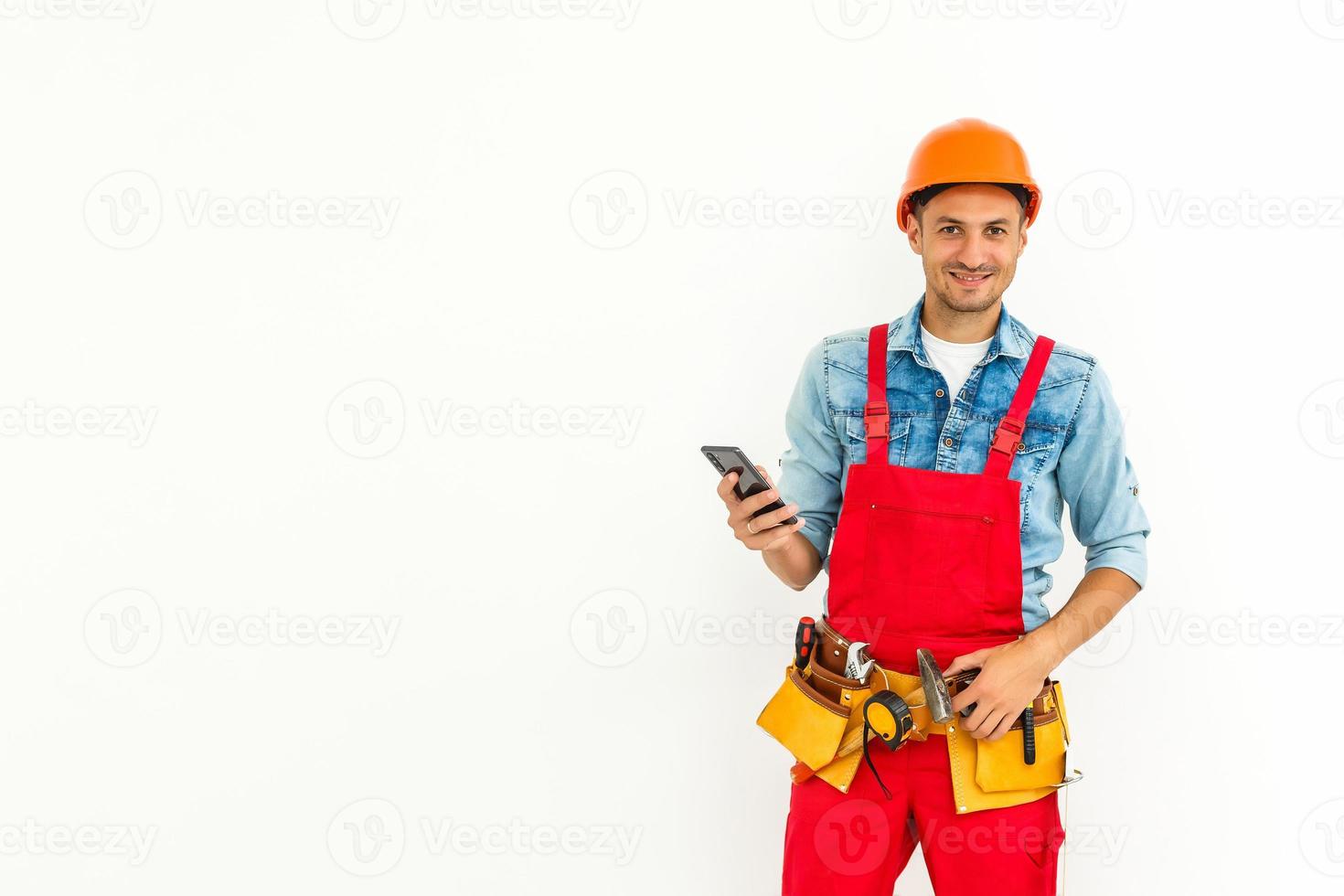 profession, construction et bâtiment - travailleur masculin souriant heureux ou constructeur en casque et dans l'ensemble avec smartphone sur fond blanc photo