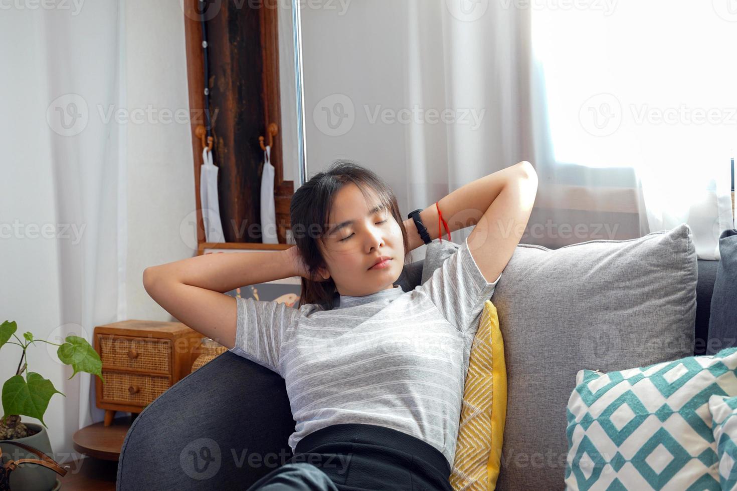 une femme asiatique ferme les yeux et s'appuie sur le canapé. elle posa sa tête sur ses mains entrelacées. pour vous reposer confortablement pendant votre temps libre. concept se détendre après le travail, se détendre, coin confortable, coin calme, sieste photo