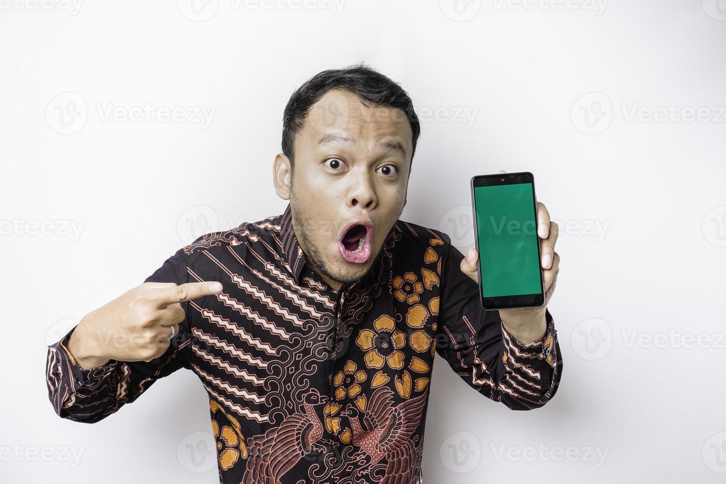 homme asiatique choqué portant une chemise batik et montrant un écran vert sur son téléphone, isolé sur fond blanc photo