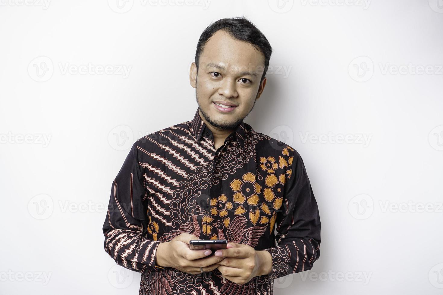 un portrait d'un homme asiatique heureux portant une chemise batik et tenant son téléphone, isolé sur fond blanc photo