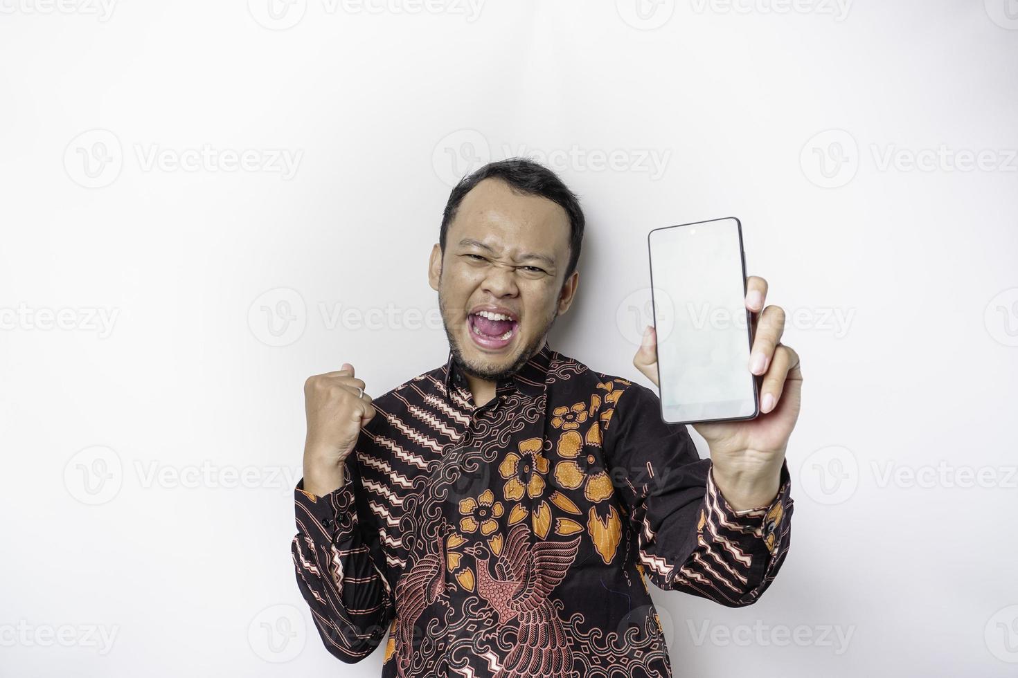 un jeune homme asiatique heureux portant une chemise batik montrant une expression réussie montrant l'espace de copie sur son téléphone isolé par fond blanc photo