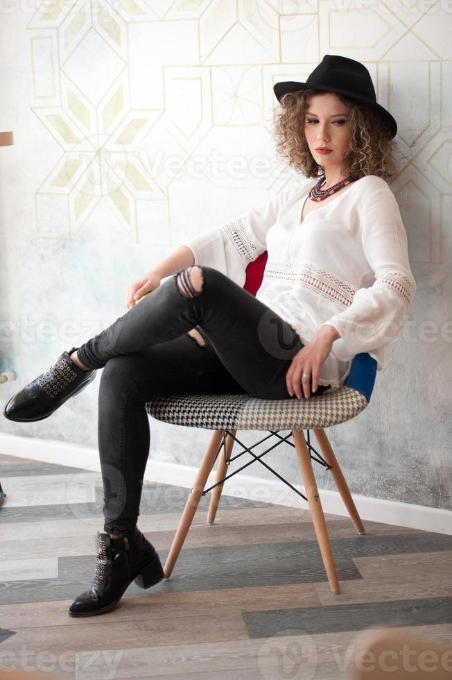 heureuse fille brune avec un chapeau noir assis à l'intérieur du bar, buvant un verre de jus vert froid tout en souriant au loin. jeune jolie femme sur le banc buvant du jus portant une chemise blanche photo