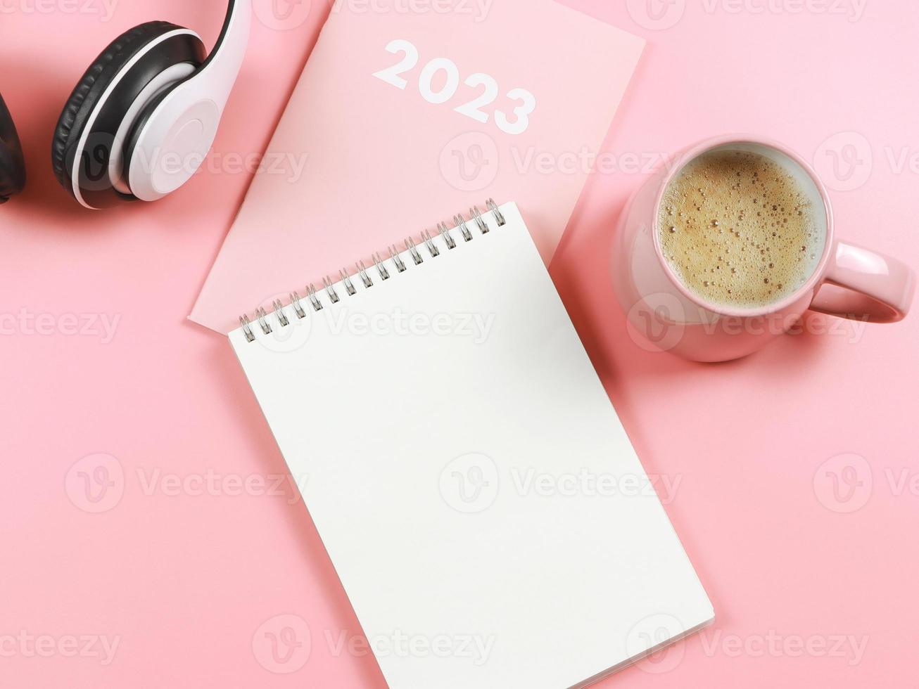 mise à plat d'un carnet de notes en papier vierge sur un agenda ou un planificateur rose 2023, un casque et une tasse de café rose sur fond rose avec espace de copie. photo