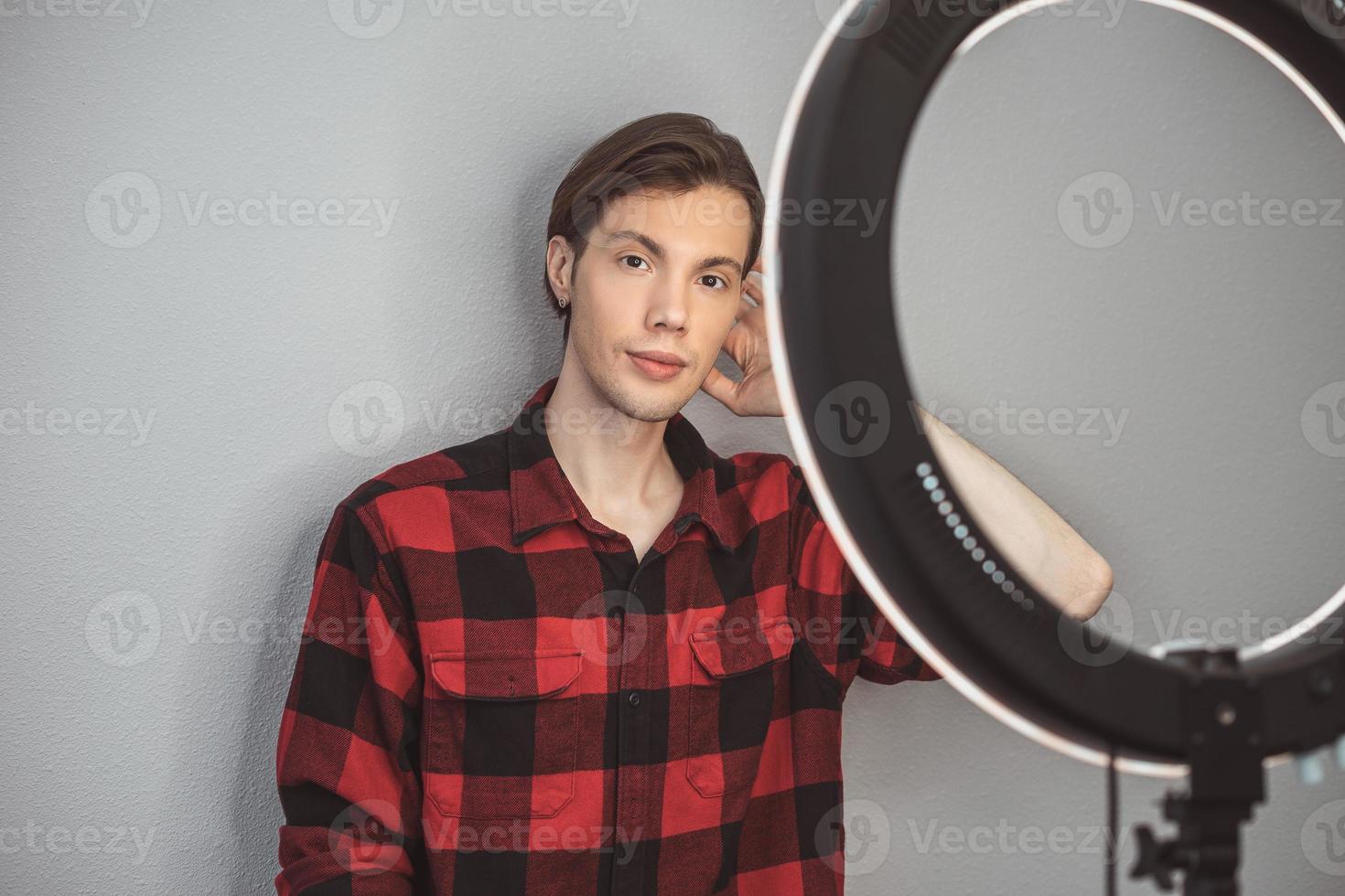 jeune homme coiffeur en chemise à carreaux rouge avec lampe annulaire sur le fond de mur gris photo