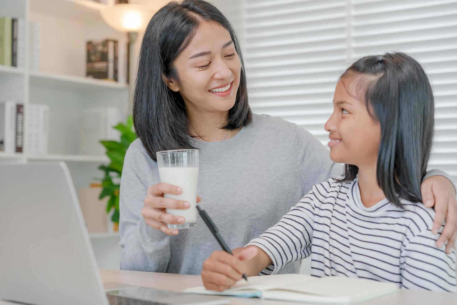 homeschool asiatique jeune petite fille apprend, lit et fait ses devoirs avec l'aide d'une mère aimable, enseigne et encourage. maman passe un verre de lait à sa fille. fille heureuse d'étudier et d'éduquer avec maman photo