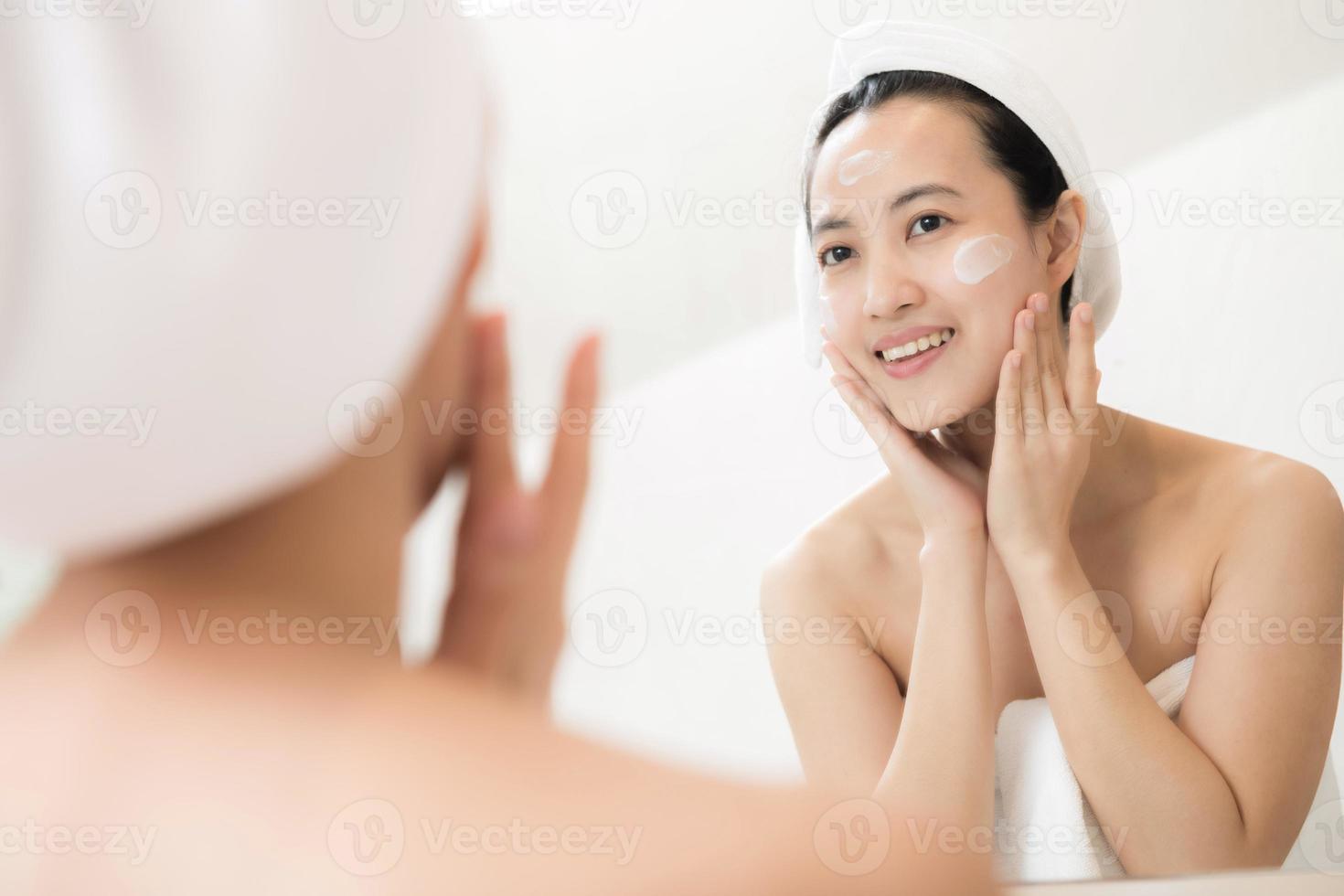 heureuse jeune femme asiatique appliquant des lotions pour le visage tout en portant une serviette et en touchant son visage dans la salle de bain photo