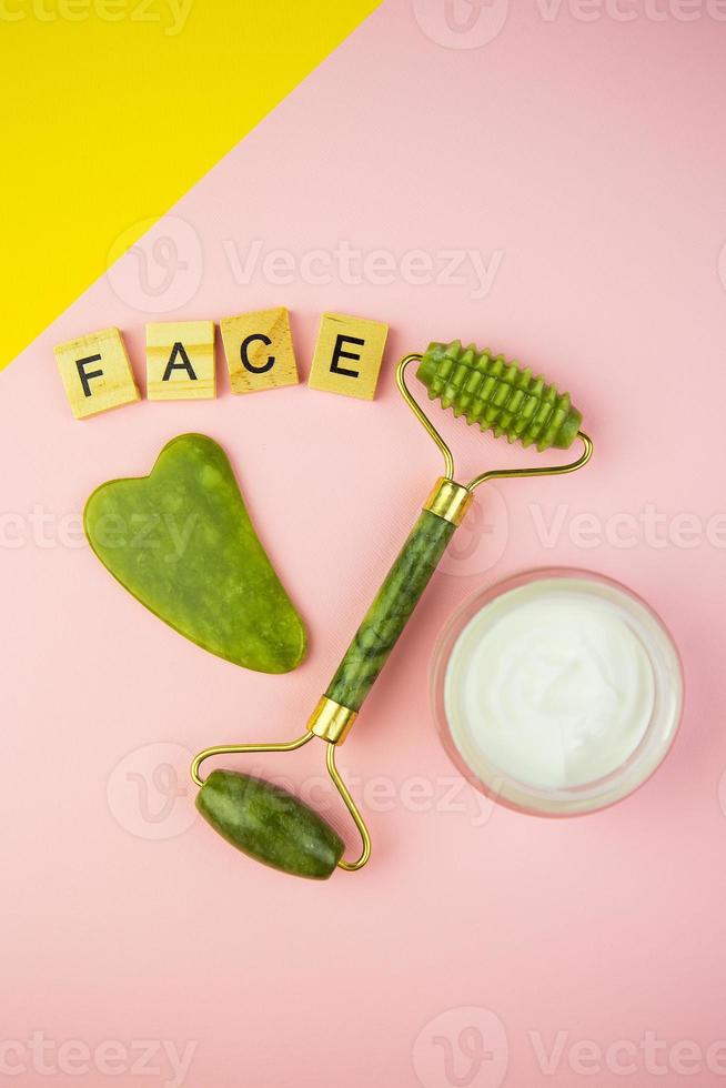 outils de massage facial vert gua sha. rouleau en jade quartz vert sur fond rose-jaune. pot de crème, lettrage facial en lettres de bois. photo