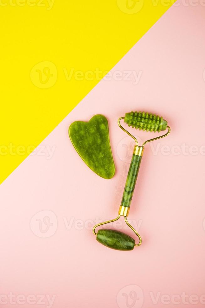 outils de massage facial vert gua sha. rouleau de jade quartz vert sur fond rose et jaune. soins anti-âge, liftants et tonifiants à domicile. espace de copie. photo