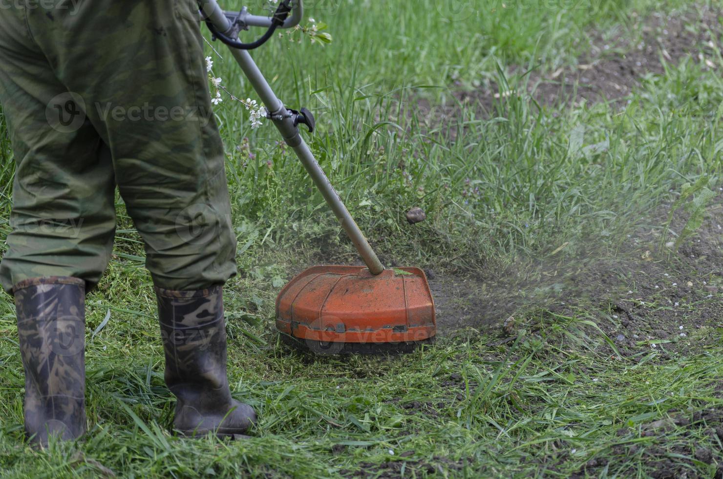 un gros plan d'un travailleur portant des vêtements de protection, des gants, des bottes en caoutchouc avec une tondeuse à essence sur la pelouse avant. un homme tond l'herbe avec des pissenlits par une chaude journée de printemps ensoleillée. photo