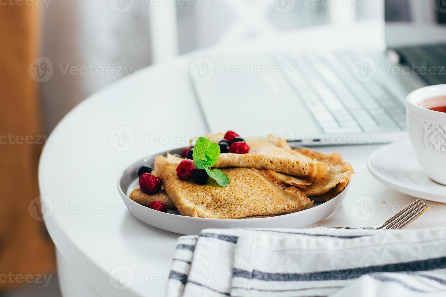 table de bureau avec ordinateur portable, crêpes fraîches, tasse de thé et pot de miel. photo