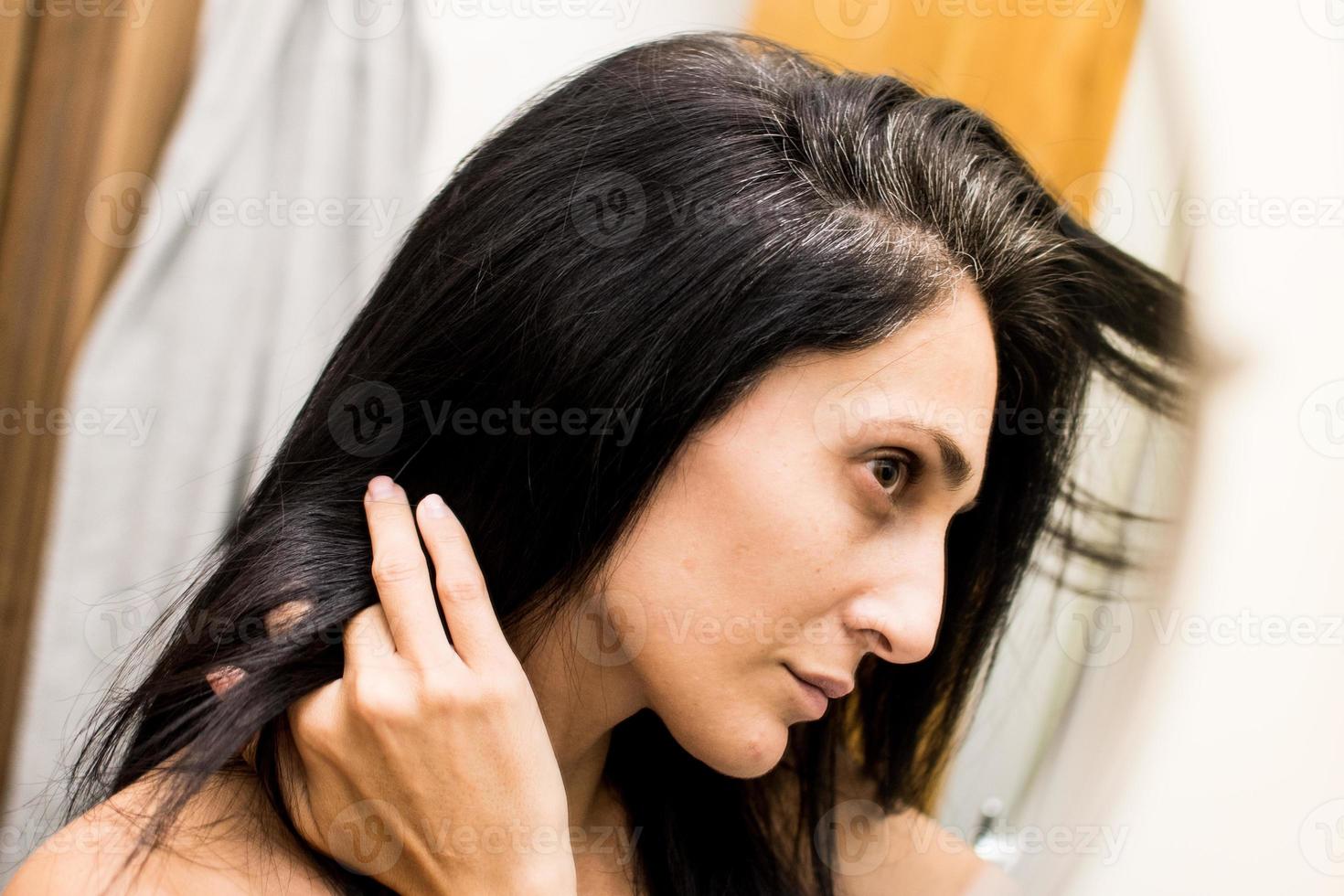 portrait d'une belle jeune femme examinant son cuir chevelu et ses cheveux devant le miroir, les racines des cheveux, la couleur, les cheveux gris, la perte de cheveux ou le problème du cuir chevelu sec photo