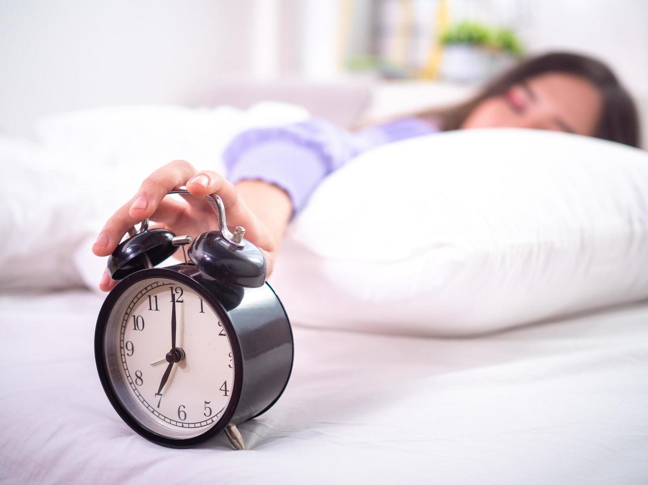 une fille endormie au lit, les yeux fermés, tend la main au réveil à la maison. bonjour un jour paresseux photo