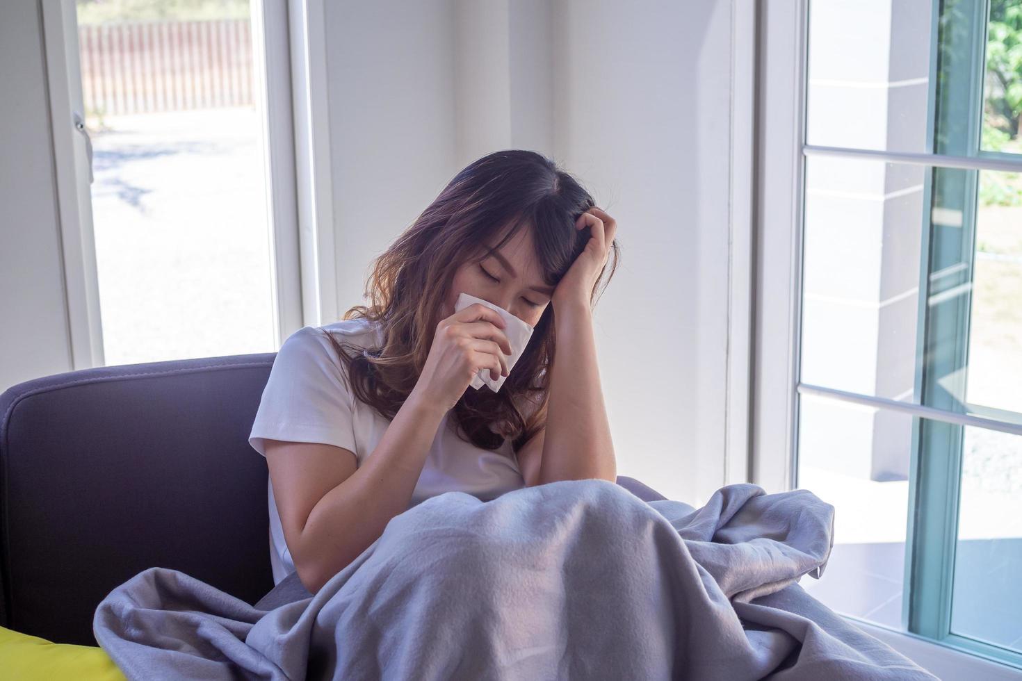 une femme aux cheveux longs assise sur le canapé souffre de grippe, de toux et d'éternuements. assis dans une couverture à cause d'une forte fièvre et se couvrir le nez avec du papier de soie parce qu'il éternue tout le temps. photo