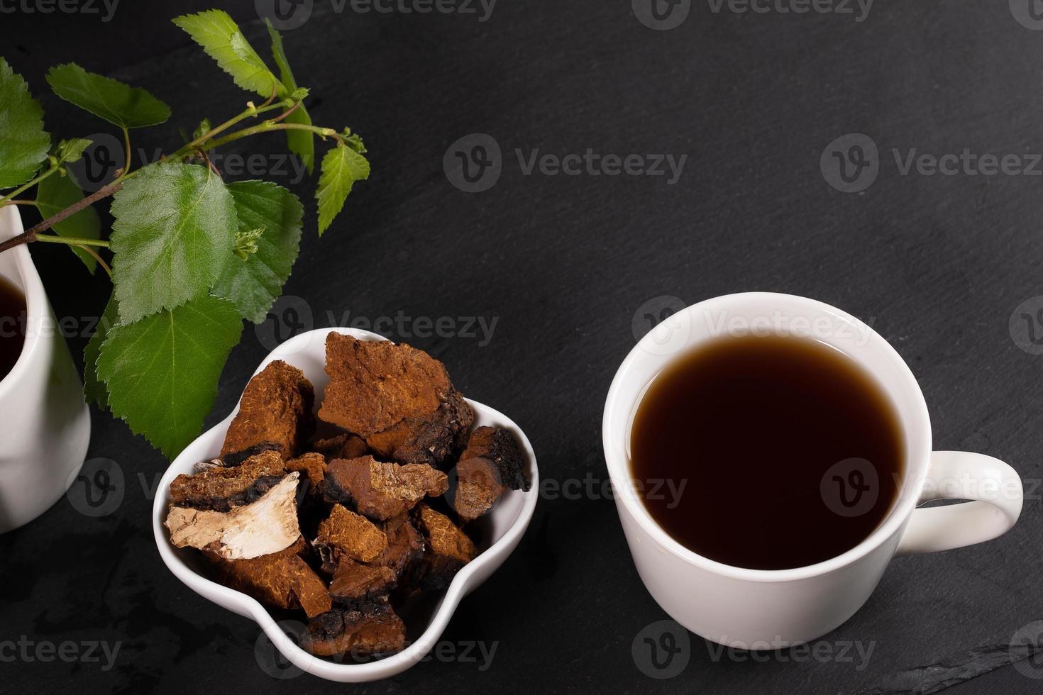 tasse en céramique blanche d'infusion curative de chaga aux champignons de bouleau, bol avec morceaux de chaga et feuilles de bouleau sur fond noir. photo