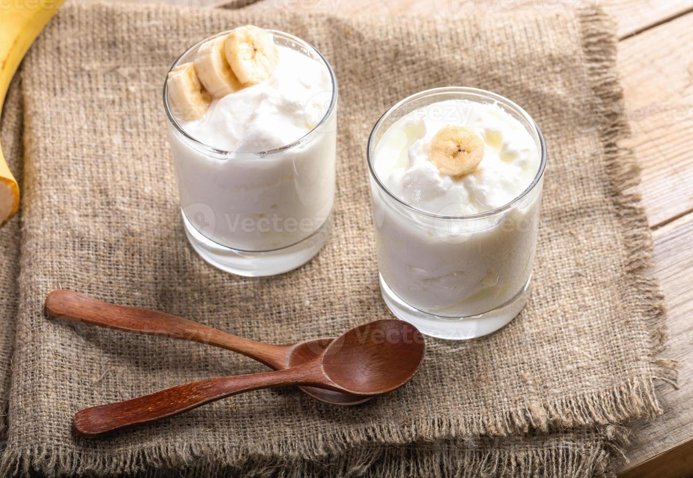 deux verres de yaourt blanc avec des tranches de banane et des cuillères en bois sur une serviette en toile de jute rugueuse. photo