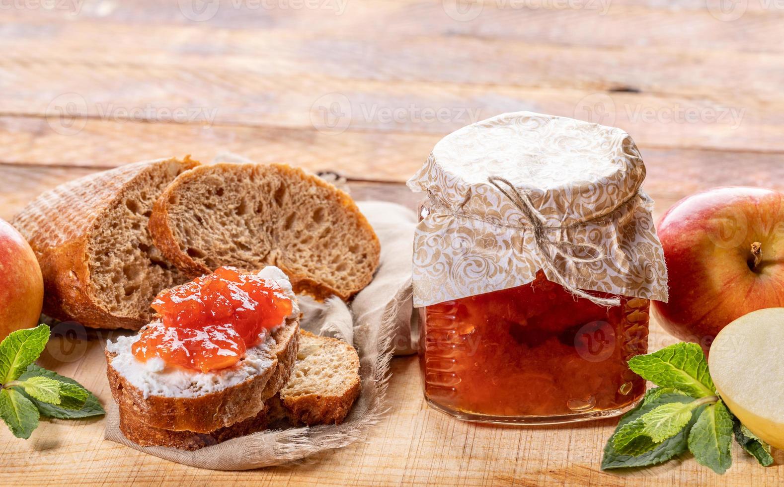 petit bocal en verre avec confiture de pomme, pain, enduit de confiture de pomme, pommes et menthe sur fond bois. photo