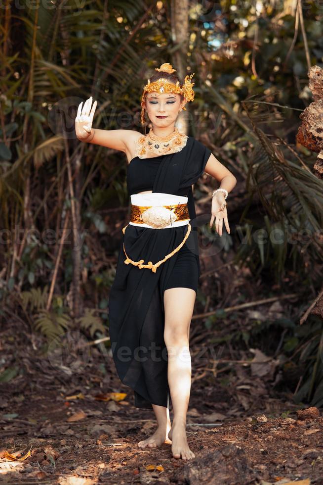 pose de danse javanaise dans un débardeur noir et une jupe noire avec une couronne dorée et des accessoires dorés sur son corps à l'intérieur de la jungle photo