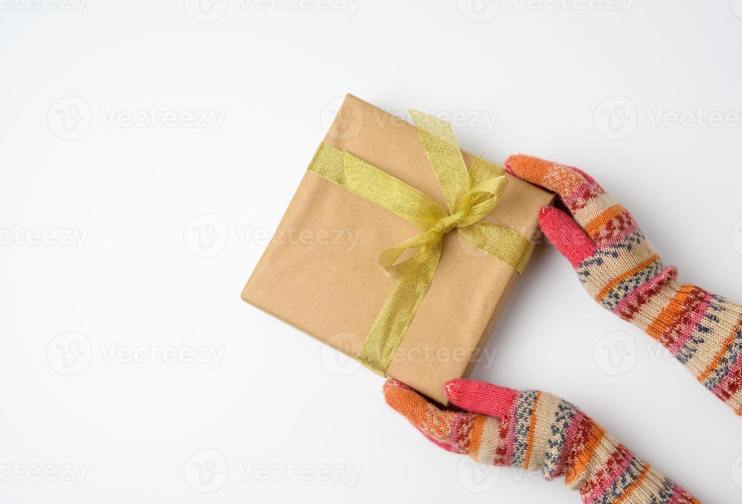 les mains des femmes dans des mitaines tricotées tiennent une boîte carrée enveloppée de papier brun et attachée avec un ruban doré photo