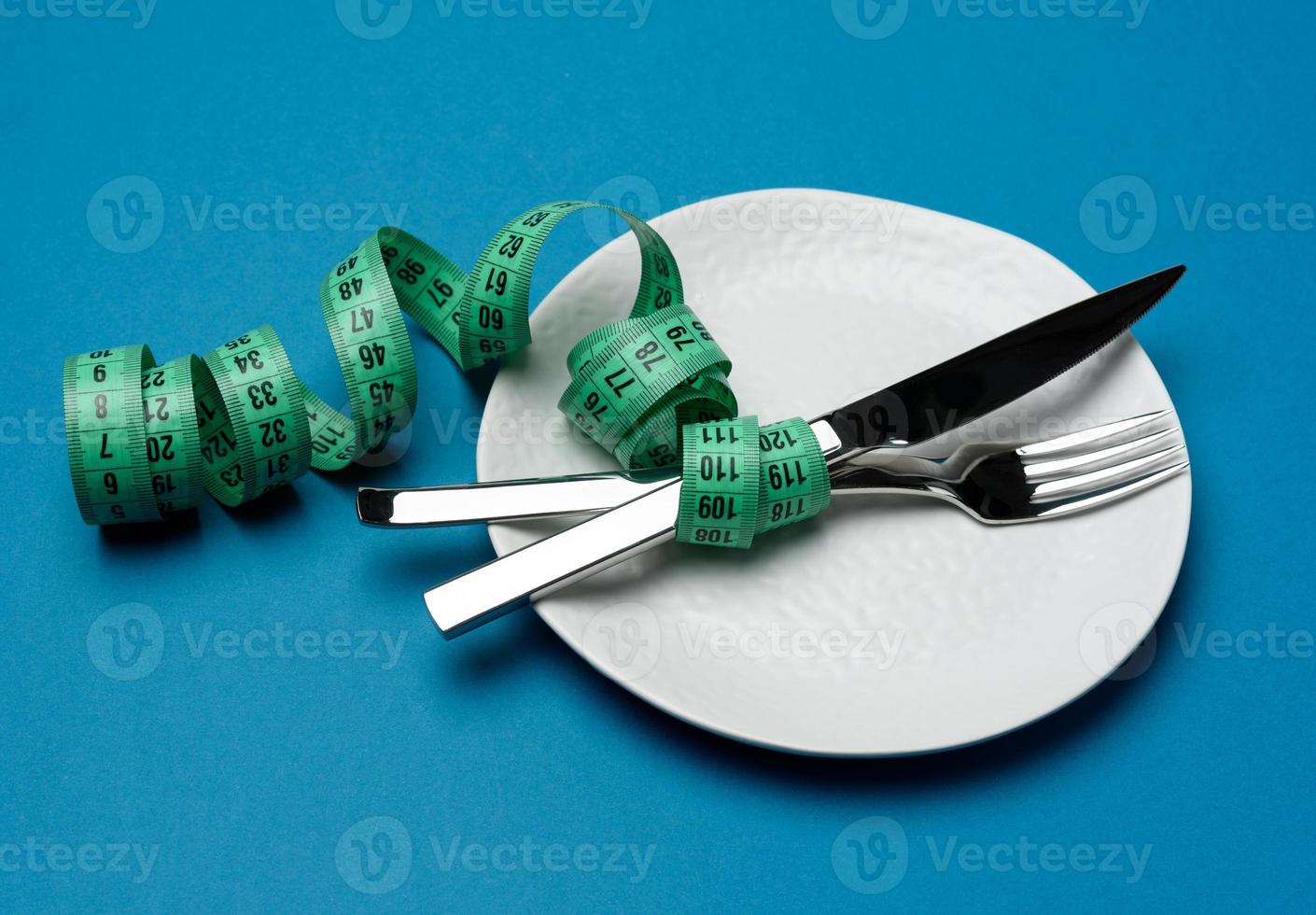 assiette ronde blanche et fourchette et couteau enveloppés dans un ruban à mesurer vert sur fond bleu photo