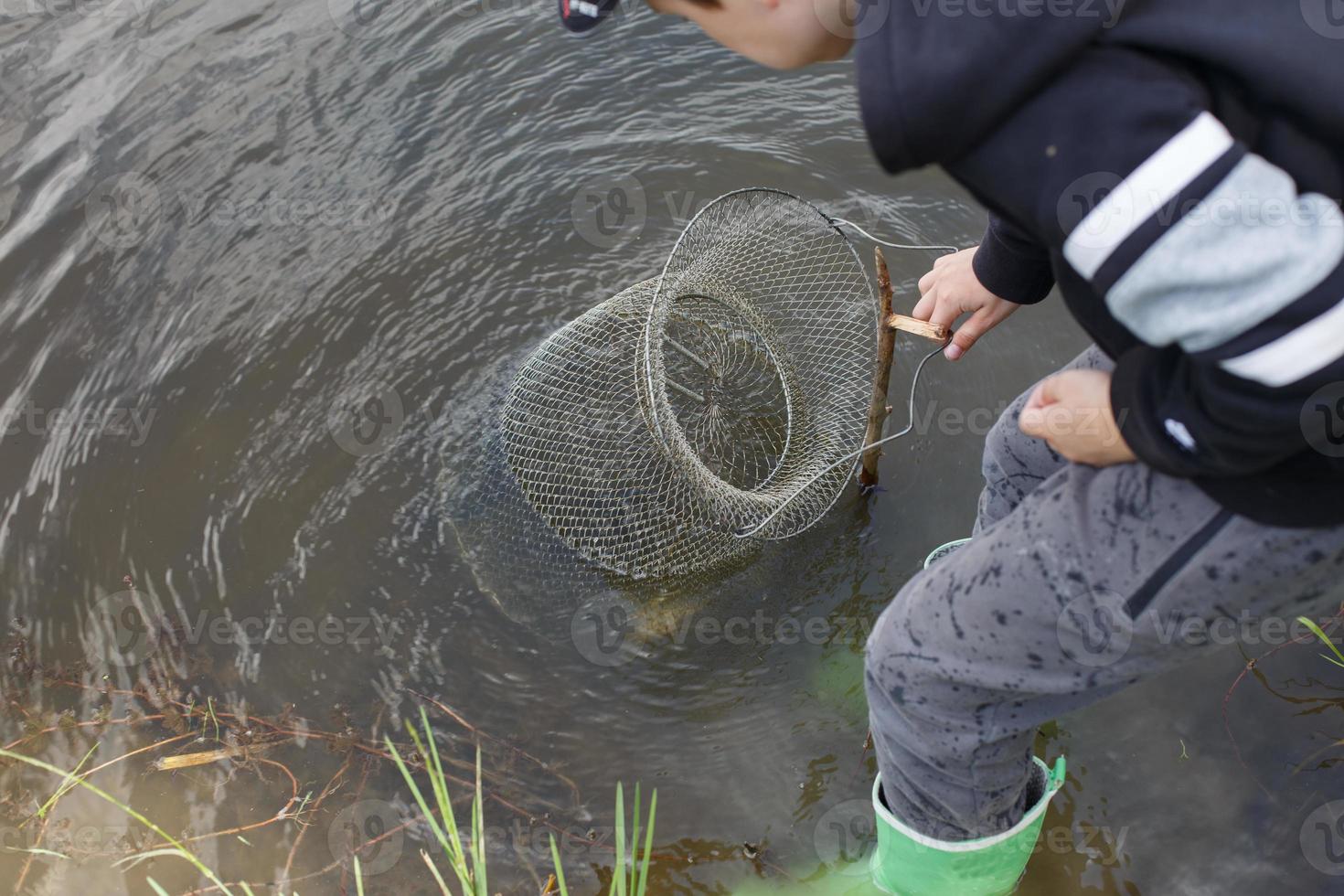 garçon enfant pêcheur soulève un filet de pêche. une cage en treillis métallique est installée dans l'eau de la rivière près du rivage photo