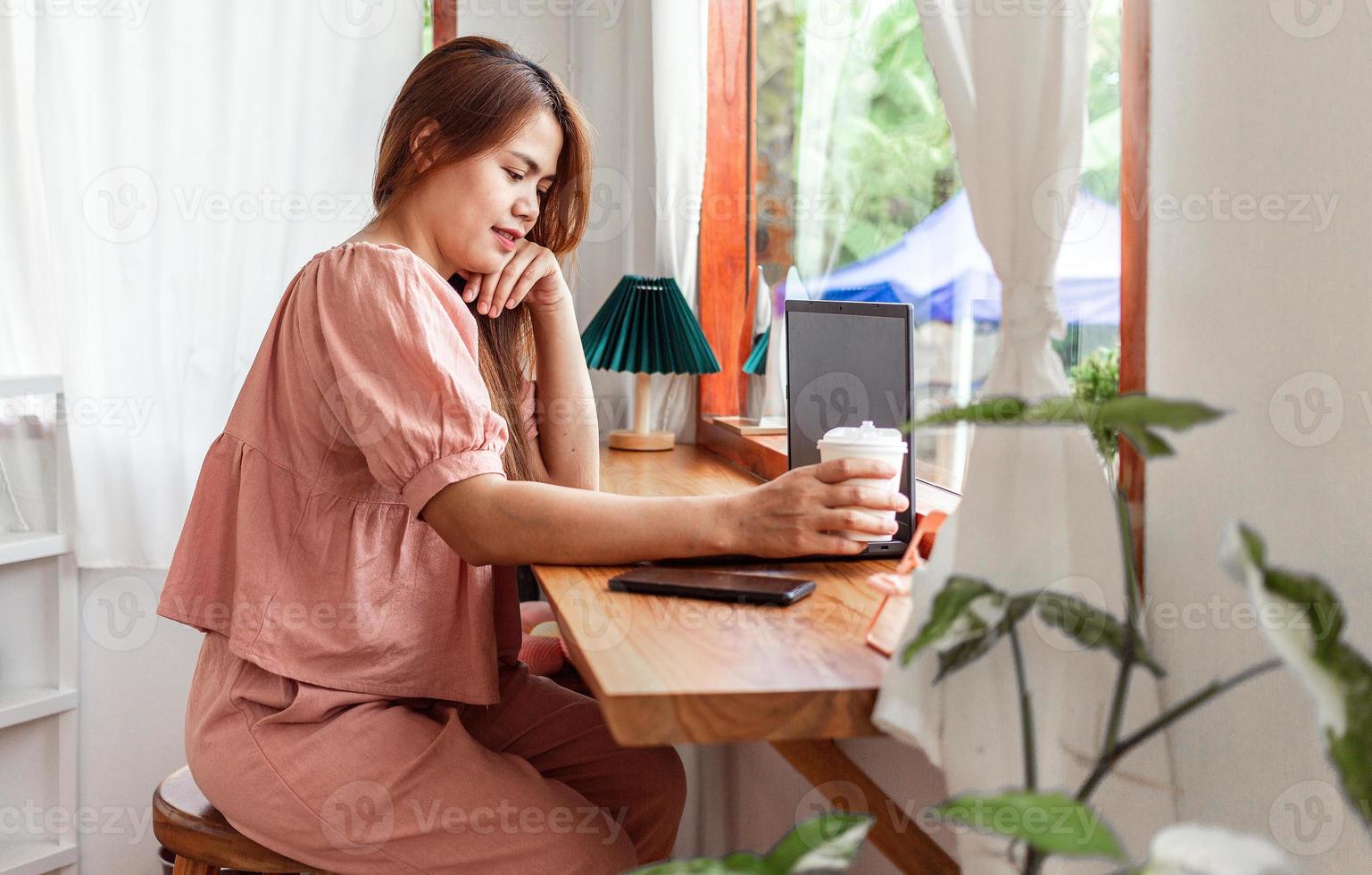 une femme heureuse dans un café utilisant un ordinateur portable à la main et une tasse de café en papier. jeune femme blanche aux cheveux longs assise dans un café occupée à travailler sur son ordinateur portable. photo