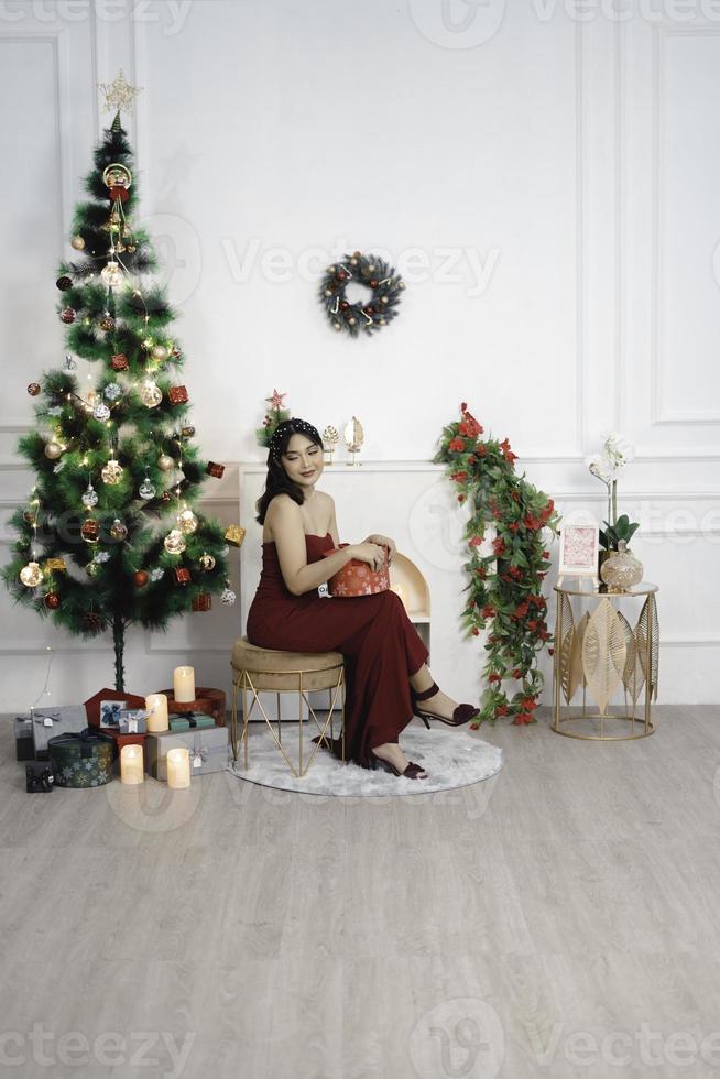 portrait de jolie jeune fille confortable s'asseoir tenant le cadeau de noël, souriant porter une robe rouge dans le salon de noël décoré à l'intérieur photo