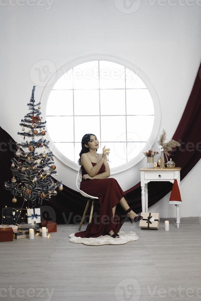 portrait d'une jolie jeune fille confortable s'asseoir et boire du vin, porter une robe rouge dans un salon de noël décoré à l'intérieur photo