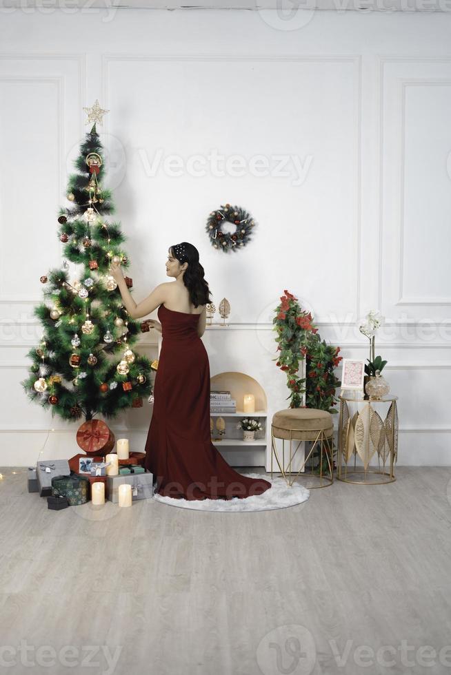 portrait d'une jolie jeune fille décorant un sapin de noël, souriante vêtue d'une robe rouge dans un salon de noël décoré à l'intérieur photo