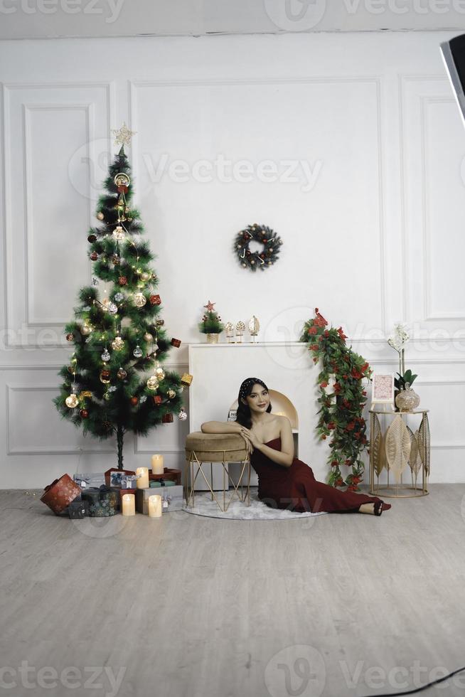 portrait de jolie jeune fille confortable s'asseoir sur le sol, souriant porter une robe rouge dans le salon de noël décoré à l'intérieur photo