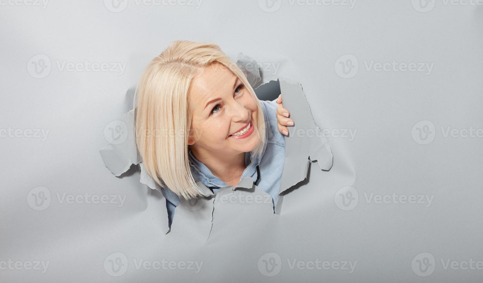 femme joyeuse charismatique avec une coiffure blonde, à l'air ludique, porte un sweat à capuche à la mode, se tient dans un trou de papier déchiré, remarque drôlement agréable. fond gris. photo