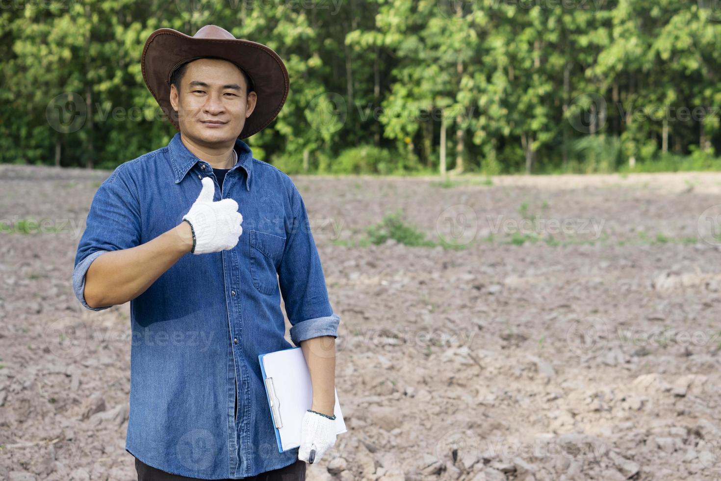 l'agronome asiatique porte un chapeau, une chemise bleue, tient un presse-papiers, le pouce levé, se tient sur les terres agricoles. concept, recherche agricole, enquête et étude pour développer la qualité du sol pour la culture des cultures. photo
