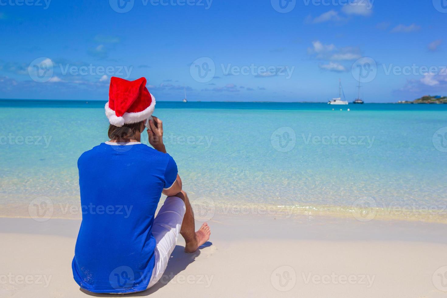 vue arrière du jeune homme en bonnet de Noel avec téléphone sur la plage tropicale des Caraïbes photo