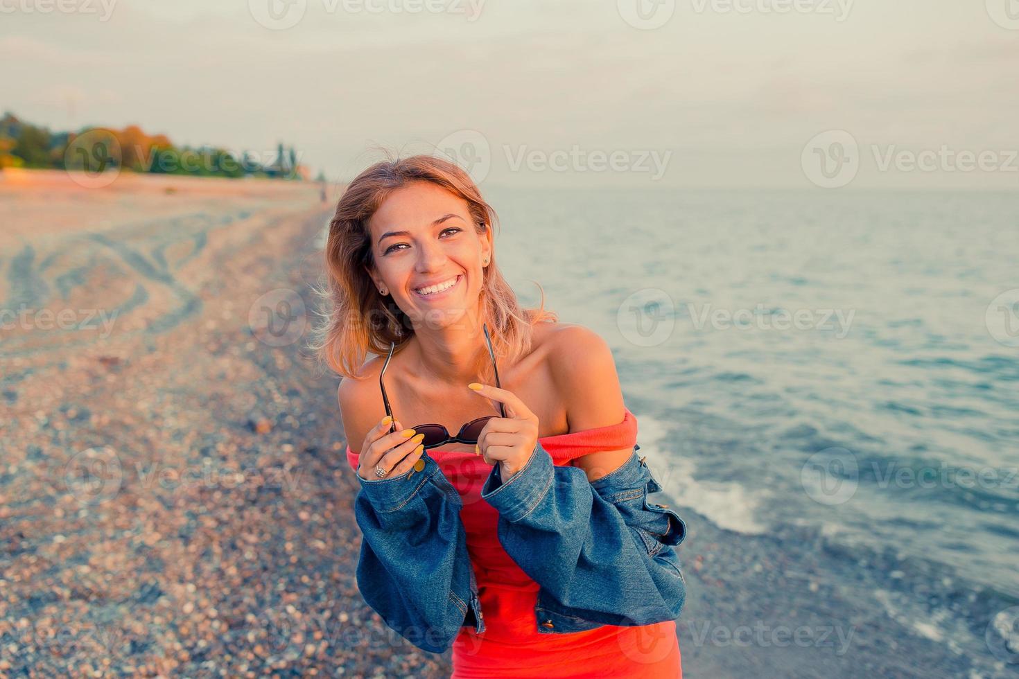 portrait de mode en plein air d'une fille élégante portant des lunettes de soleil à la mode et une veste en jean sur la plage. mains en l'air, filtre rétro, taches de soleil photo