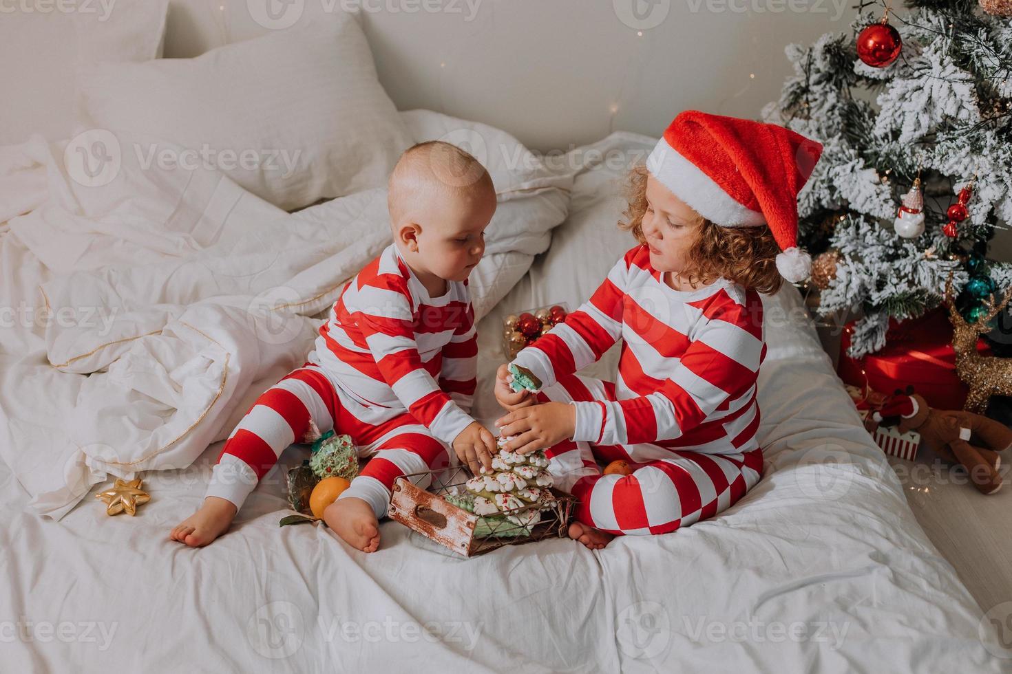 les enfants en pyjama rouge et blanc mangent des bonbons de noël assis dans leur lit. frère et sœur, garçon et fille partagent des cadeaux. Matin de noël. mode de vie. espace pour le texte. photo de haute qualité