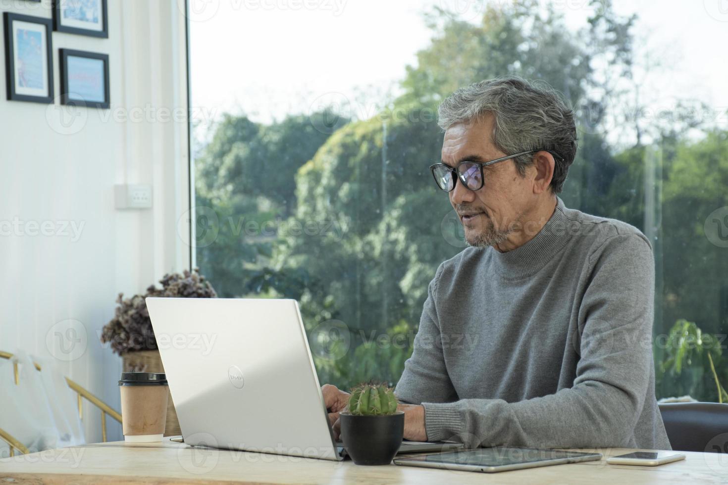 Portrait d'un homme asiatique senior âgé assis près d'une fenêtre en verre le matin pour travailler à domicile et vérifier ses affaires sur son ordinateur portable sur la table avec une mise au point sérieuse, douce et sélective. photo