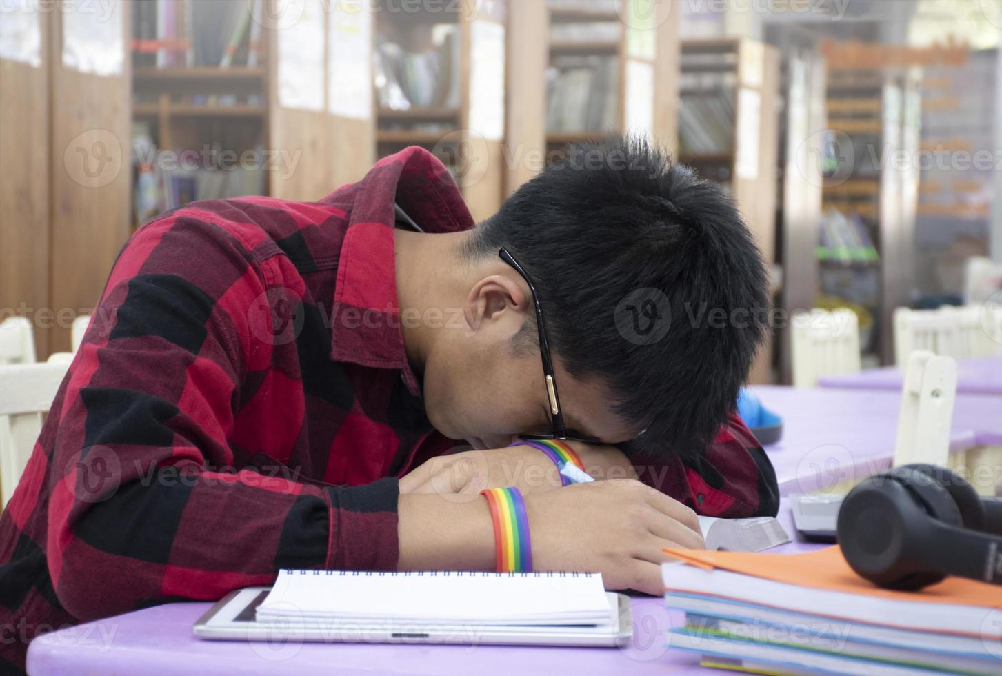 jeune garçon asiatique porte un bracelet arc-en-ciel, assis dans la bibliothèque et faisant une sieste sur la table tout en faisant son dur travail de projet scolaire, concept pour élever les activités de la vie quotidienne des adolescents et des personnes lgbt. photo